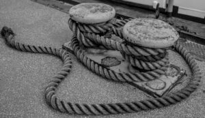 Schwarz-weiß-Foto zweier massiver Festmacher-Poller auf einem Schiff, um die dicke, verwitterte Trosse mehrfach geschlungen ist. Das Seil liegt locker auf dem Boden und bildet weiche Kurven. Die Oberfläche der Poller zeigt deutliche Gebrauchsspuren.