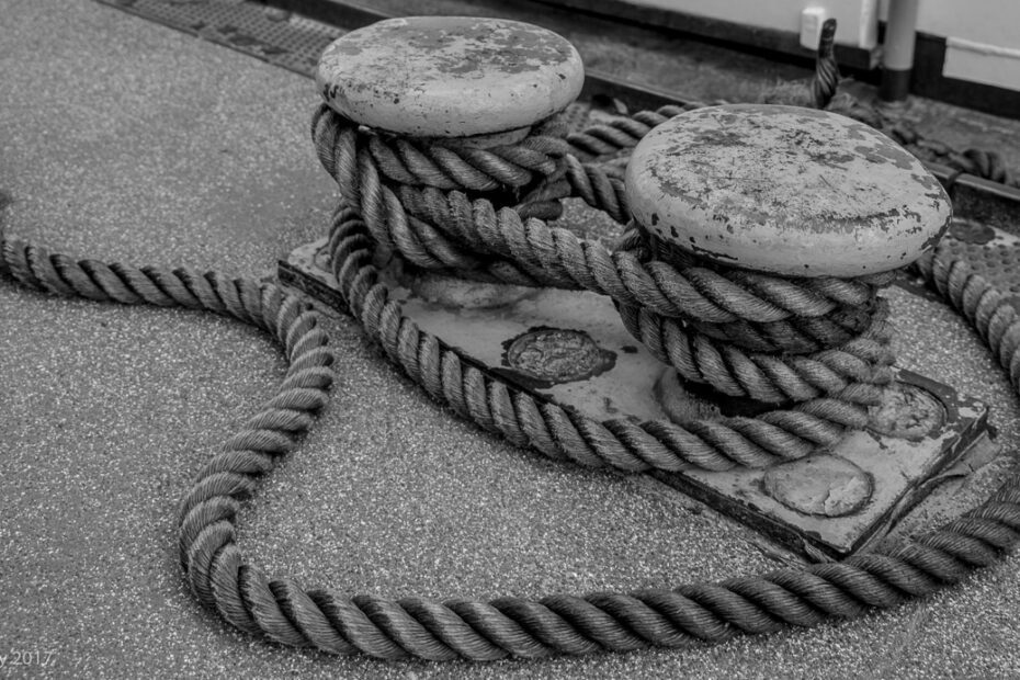 Schwarz-weiß-Foto zweier massiver Festmacher-Poller auf einem Schiff, um die dicke, verwitterte Trosse mehrfach geschlungen ist. Das Seil liegt locker auf dem Boden und bildet weiche Kurven. Die Oberfläche der Poller zeigt deutliche Gebrauchsspuren.