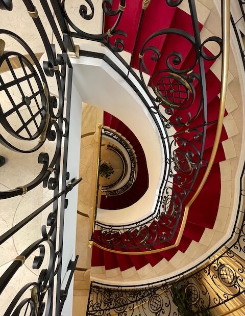 Blick von oben in ein historisches Treppenhaus mit rotem Teppich und kunstvoll geschwungenem schmiedeeisernem Geländer. Die Treppe windet sich spiralförmig nach unten, warmes Licht betont die Eleganz des alten Hotels