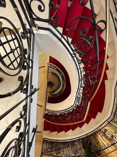Blick von oben in ein historisches Treppenhaus mit rotem Teppich und kunstvoll geschwungenem schmiedeeisernem Geländer. Die Treppe windet sich spiralförmig nach unten, warmes Licht betont die Eleganz des alten Hotels