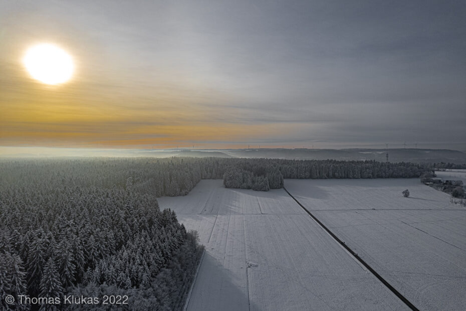Luftaufnahme einer winterlichen Landschaft im Abendlicht. Eine schmale Straße teilt das helle Schneefeld, während Nebel über den Wäldern liegt und die Sonne tief steht. Symbol für Ruhe, Klarheit und den stillen Moment zwischen Licht und Stille.